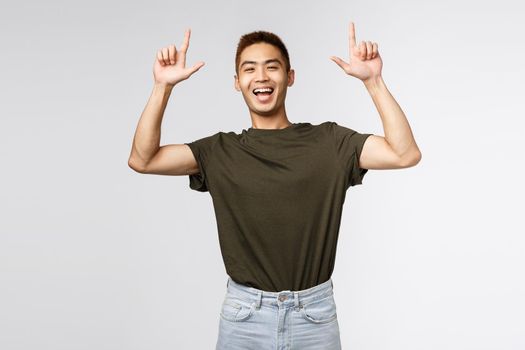 Portrait of optimistic, happy asian man in green t-shirt inviting people check-out advertisement, laughing and smiling excited, pointing fingers up, showing way to banner, grey background.