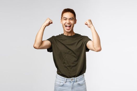 Competition, prize and people concept. Portrait of happy, triumphing young asian man raising hands up, fist pump celebrating victory, achieve success and winning, smiling upbeat, grey background.