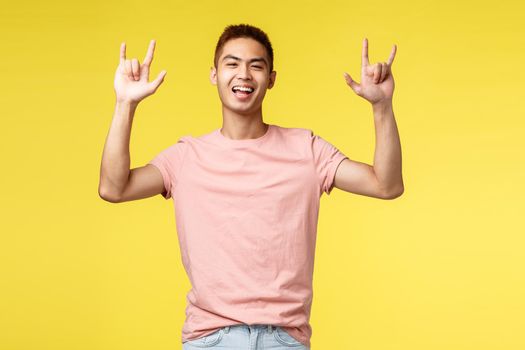 Lifestyle, education and people concept. Carefree handsome asian guy having fun, enjoying awesome summer campus party, show rock-n-roll sign and smiling broadly, partying, yellow background.