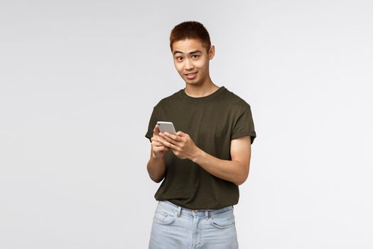 Technology, online lifestyle and communication concept. Portrait of asian surprised man, look at camera with disbelief and amusement, messaging mobile phone, texting in social media, grey background.