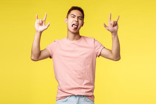 Lifestyle, education and people concept. Enthusiastic handsome and cool asian man in pink t-shirt, show tongue and rock-n-roll sign, having fun, partying, standing yellow background.