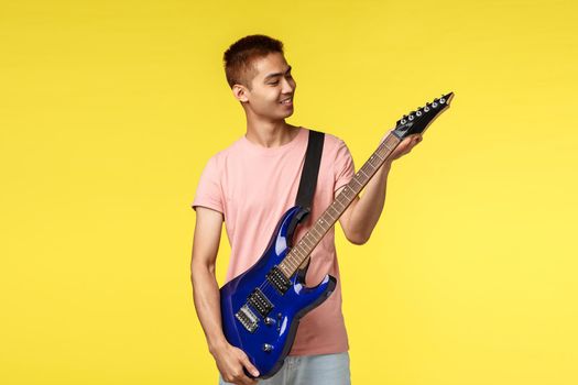 Lifestyle, leisure and youth concept. Cheerful handsome asian male student playing in band, tune electric guitar and smiling pleased, ready to jam on stage, perform new song, yellow background.