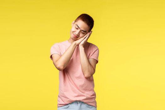 People, different emotions and lifestyle concept. Dreamy asian sleepy cute guy in pink t-shirt, dreaming about going summer vacation, lean on palms like falling asleep, smiling, yellow background.