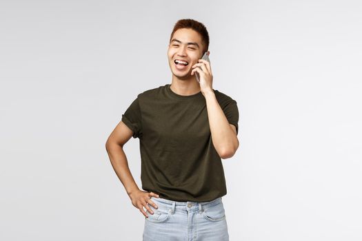 Technology, online lifestyle and communication concept. Portrait of cheerful laughing asian guy talking to friend via mobile phone, chuckle and smiling as looking away casually, grey background.