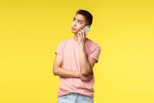 Technology, communication and lifestyle concept. Portrait of young asian guy waiting for his girlfriend pick-up phone, waiting on line as order food delivery, look indecisive up, yellow background.