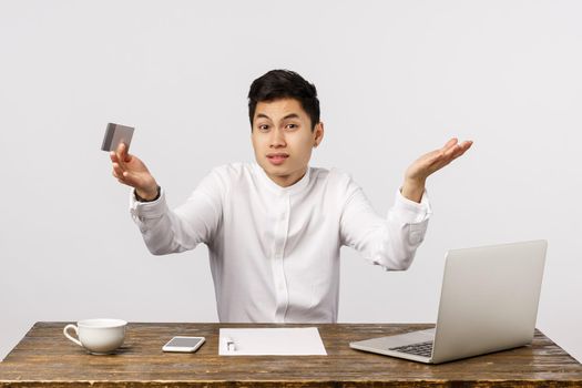 I dont know what to buy. Perplexed, unsure and confused chinese male sitting in office near laptop, documents, holding credit card, shrugging trying decide what order online, white background.