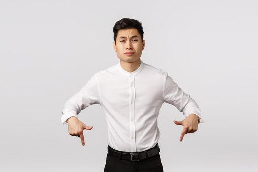 Skeptical arrogant asian businessman in white shirt, pants, pointing down look camera unsatisfied and unimpressed, sulking, grimacing careless, express dislike or scorn, white background.