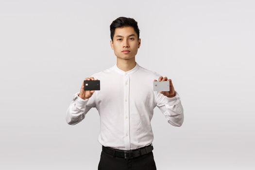 Pick your color. Serious handsome and stylish asian businessman holding two credit cards give customer choice, promote new bank finance service, advice payment variants, standing white background.