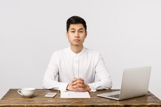 Lets get straight business. Serious-looking young asian businessman, executive having meeting with clients, sitting office desk in white shirt, with laptop, documents and tea, listening proposals.