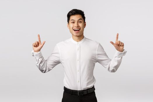 Cheerful cute asian guy celebrating birthday inviting guests see fireworks. Man rejoicing as standing in suit, pointing up, smiling happy and entertained, found product he liked, white background.