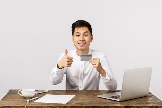 Satisfied good-looking asian office worker, manager sitting desk with documents, laptop and cup coffee, holding credit card, show thumbs-up and smiling, recommend buy online and use bank service.