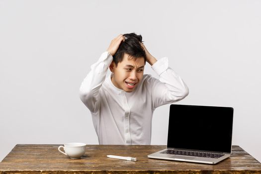 Alarmed and panicking chinese guy pulling hair from head sitting troubled and distressed, having huge problem, staring at laptop with anxious, nervous expression, sitting office white background.