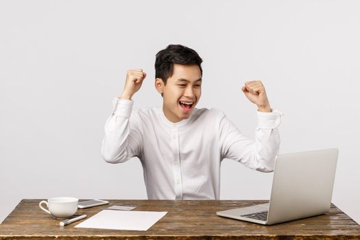 Celebration, success and finance concept. Cheerful young asian man rejoicing, raising clenched fists in hooray, yes gesture, sitting desk, looking laptop, watching online live stream game, triumphing.