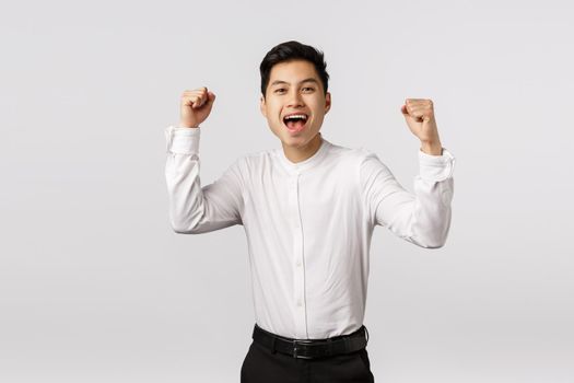 Excited, rejoicing young asian successful businessman celebrating win, become champion, achieve goal, fist pump and yelling yes as feeling pleased, team won, overjoy over victory, white background.