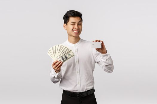 Relax bank got you covered. Cheeky and relaxed, cheerful successful asian businessman wink camera and smiling, show money and credit card, advice you put cash deposit, standing white background.