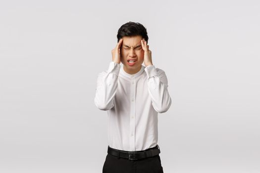 Come on need think. Tensed and distressed asian young man suffering big headache, close eyes, frowning and grimacing from pain, rubbing temples, have headache, need painkillers, white background.