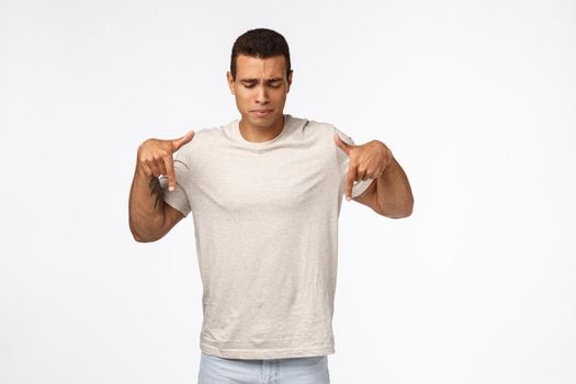 Upset, gloomy and depressed young hispanic man in t-shirt, frowning sighing sad, pointing and looking down distressed, express pity or shame of missing chance, standing frustrated white background.