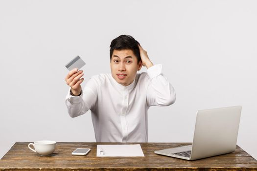 Awkward handsome chinese guy sitting office desk with laptop, documents and tea, showing credit card, scratching head worried, apologizing dont have money pay, white background.