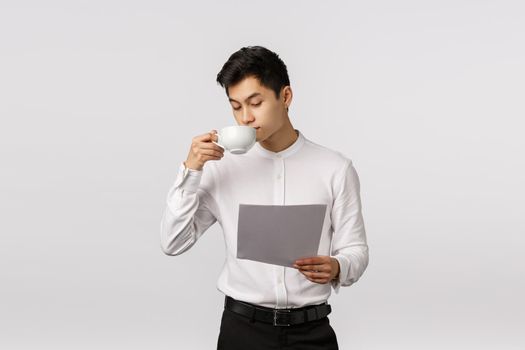 Finance, economy and business concept. Stylish and elegant, handsome asian businessman drink coffee from cup and reading documents, boss work in office study employee report, white background.