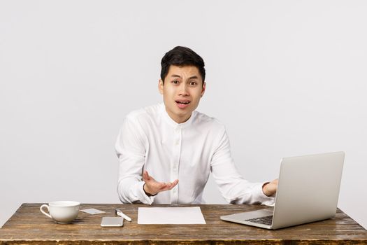 Frustrated and upset, distressed young asian office manager male, sitting table and spread hands sideways dismay, complaining, having failure, bad results, feeling regret and disappointment.