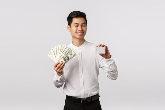 Finance, economy and business concept. Handsome asian businessman in formal outfit, holding cash and credit card, looking at banking payment method with pleased smile, spend money.