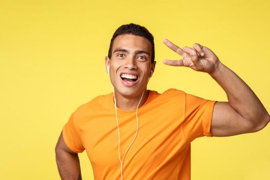 Cheerful, lucky smiling man in orange t-shirt, listen motivational music in headphones, showing peace or victory sign, grinning optimistic, enjoy favorite songs, standing yellow background upbeat.