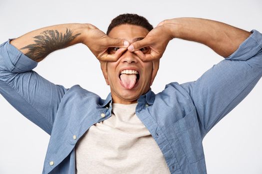 Happiness, holidays and people concept. Joyful good-looking young hispanic guy in blue shirt over t-shirt, showing tongue happily mimicking superhero, place hands over eyes like mask or glasses.