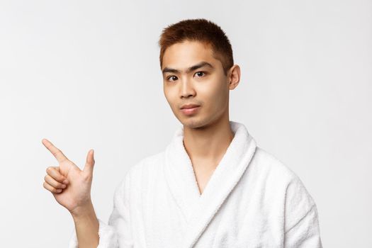 Beauty, spa and leisure concept. Portrait of young asian man inviting friends to hot springs, pointing fingers left and looking camera, show advertisement, wearing bathrobe, white background.