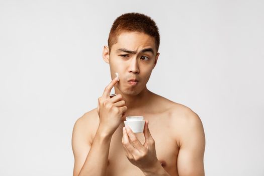 Beauty, people and leisure concept. Handsome asian naked man standing in bathroom, grimacing as looking at mirror and applying daily skincare routine cream, standing white background.