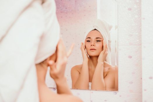 Young beautiful woman using a moisturizing facial mask after taking a bath. Pretty attractive girl in a towel on her head stands in front of a mirror in a home bathroom. Daily hygiene and skin care.