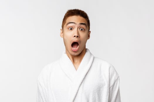 Beauty, spa and leisure concept. Portrait of amazed, startled asian man open mouth, hear amazing news, wearing bathrobe, talking to friend, head something interesting, white background.