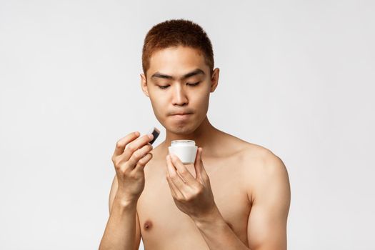 Beauty, people and leisure concept. Portrait of curious asian man with naked torso open girlfriend facial cream, use cosmetic product for skincare routine, standing white background.