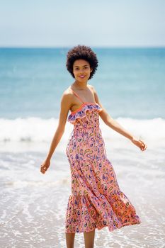 Happy black woman in summer dress, walking on the sand of a tropical beach. Girl enjoying her holiday in a coastal area.