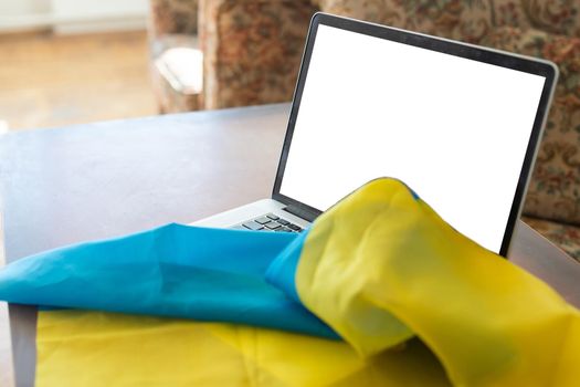 Laptop with blank screen on table and Ukrainian national flag on background, space for text