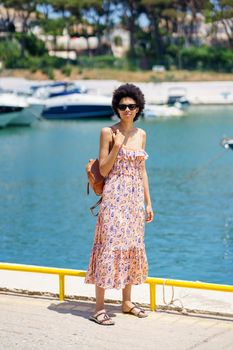 Happy black female with afro hairstyle in summer dress, walking through a harbour full of boats. Girl enjoying her holiday in a coastal area.