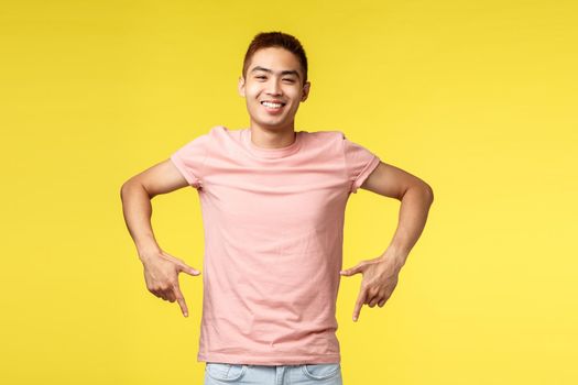 Portrait of easy-going joyful asian guy in pink t-shirt, inviting visit event, pointing fingers down, smiling pleased, recommend product, showing bottom promo, standing yellow background.