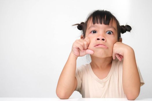 Cute dark haired little girl with angry face sitting at a table on a white background and looking at the camera.