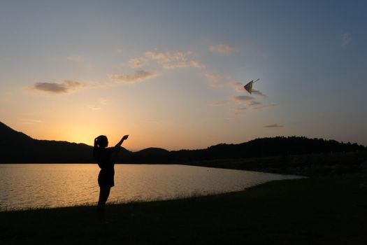 Silhouette of young woman playing a kite by the lake at sunset.