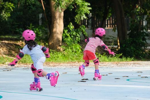 Cute Asian little girls in protective pads and safety helmet practicing roller skating in the park. Exciting outdoor activities for kids. Siblings help and support each other.