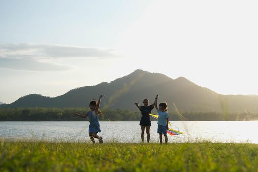 Children are flying kites while running on a meadow by the lake at sunset with their mother. Healthy summer activity for children. Funny time with family.