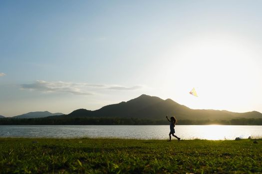 Child playing with a kite while running on a meadow by the lake at sunset. Healthy summer activity for children. Funny time with family.