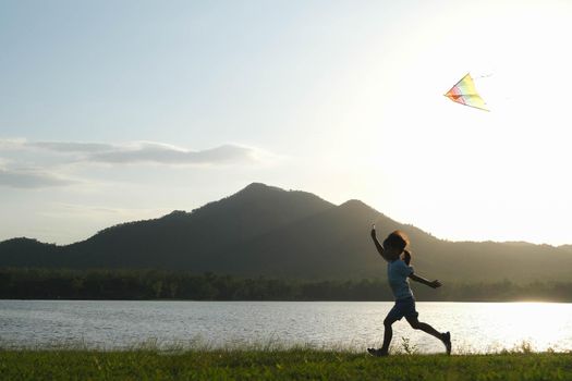 Child playing with a kite while running on a meadow by the lake at sunset. Healthy summer activity for children. Funny time with family.