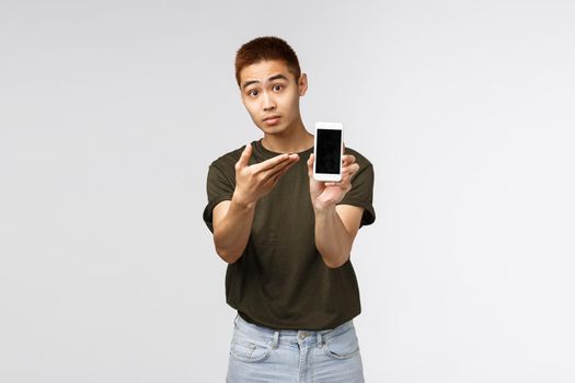 Technology, online lifestyle and communication concept. Look at this. Portrait of curious handsome asian young male asking opinion, showing smartphone display and look camera interested.