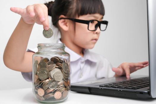 Little businesswoman puts a coin in a glass jar on a table while working with laptop in office. Children and business concept