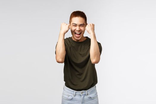 Cheerful happy, luck asian male student winning competition, achieve goal, celebrate success with beaming smile, fist pump as rejoicing and triumphing, celebrate victory over grey background.