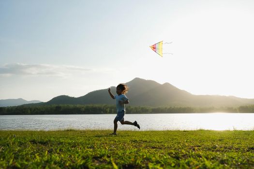 Child playing with a kite while running on a meadow by the lake at sunset. Healthy summer activity for children. Funny time with family.