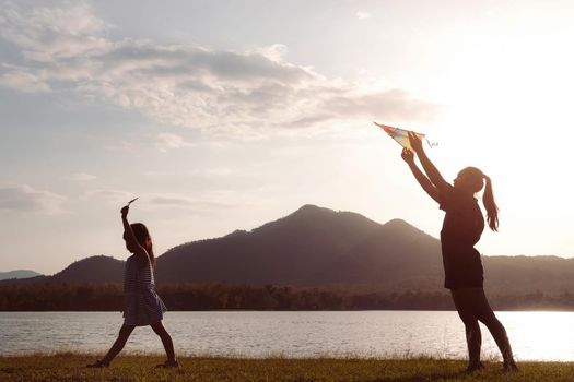 Cute little girl flying a kite while running on a meadow by the lake at sunset with her mother. Healthy summer activity for children. Funny time with family.