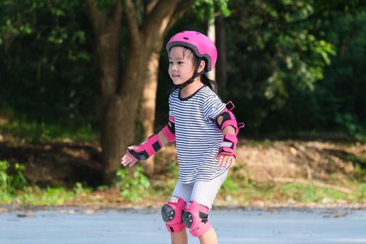 Cute Asian little girl in protective pads and safety helmet practicing roller skating in the park. Exciting outdoor activities for kids.