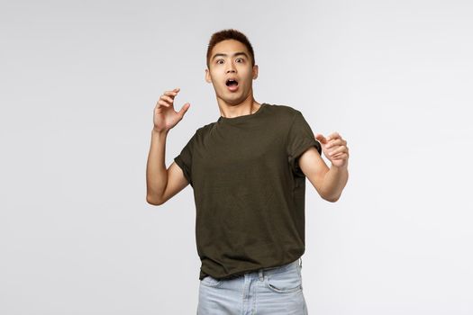 Portrait of alarmed and scared, shocked asian guy gasping, step back and raising hands up frightened, startled staring at something frightening, standing in awe over grey background.