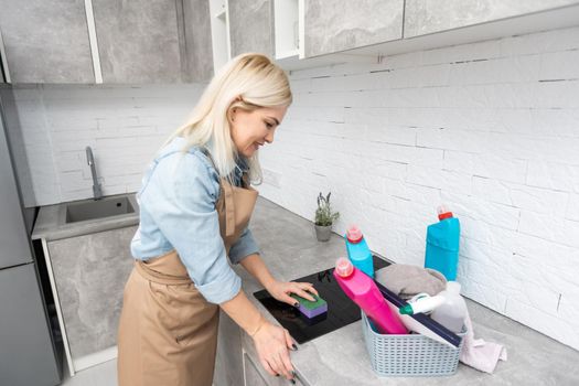 People, housework and housekeeping concept. Blonde middle aged woman with bottle of spray cleanser cleaning oven with sponge at home kitchen